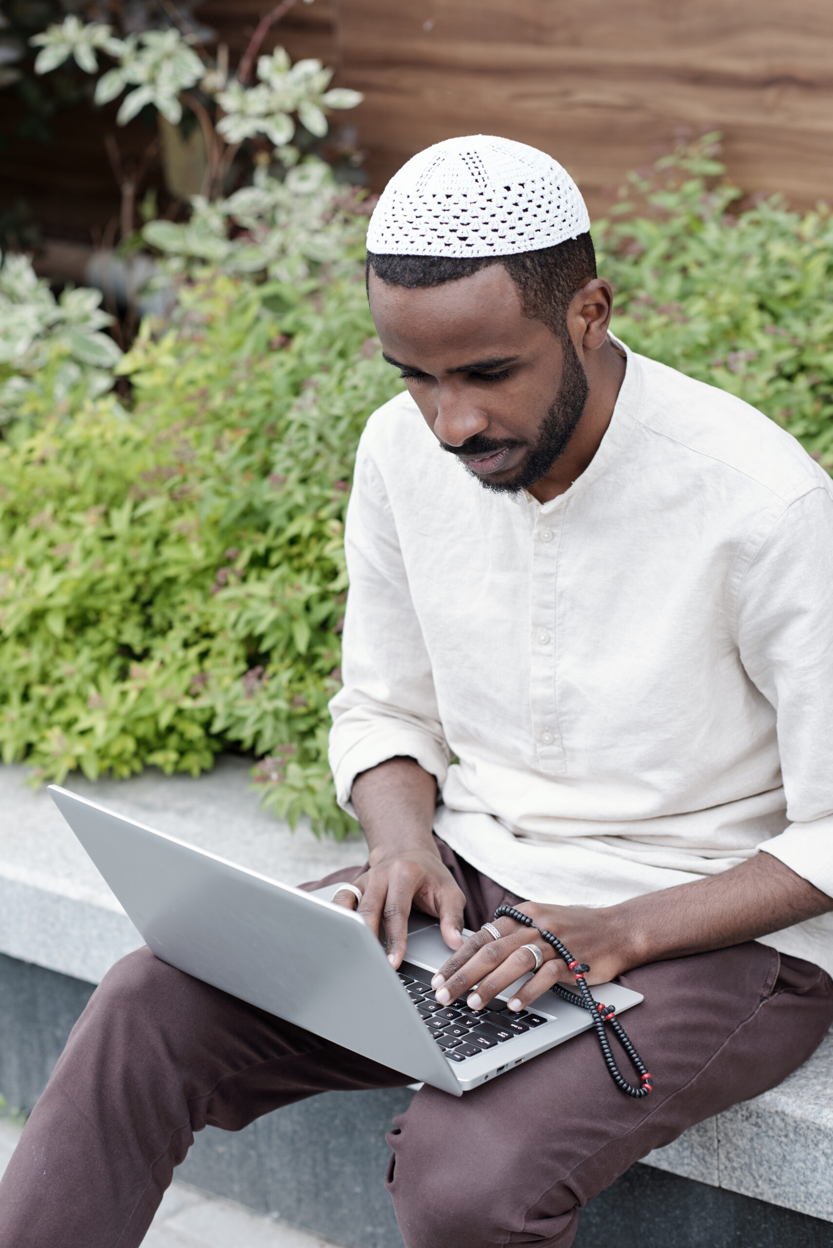 Concentrated busy young Muslim man in kufi cap sitting on curb and working with laptop